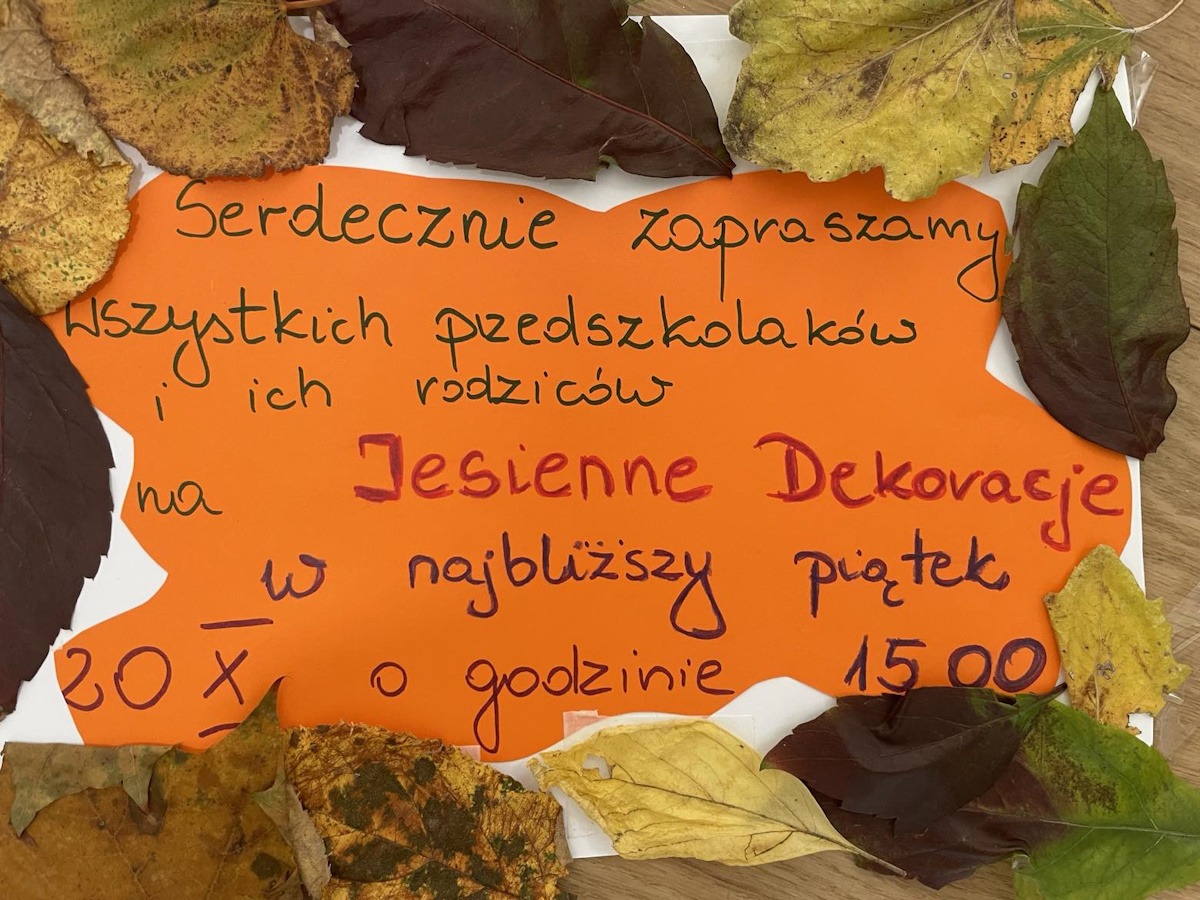 Zaproszenie na jesienne dekoracje do przedszkola przy ul. AK Żmija. Brązowe napisy na pomarańczowej kartce otoczonej jesiennymi liściami